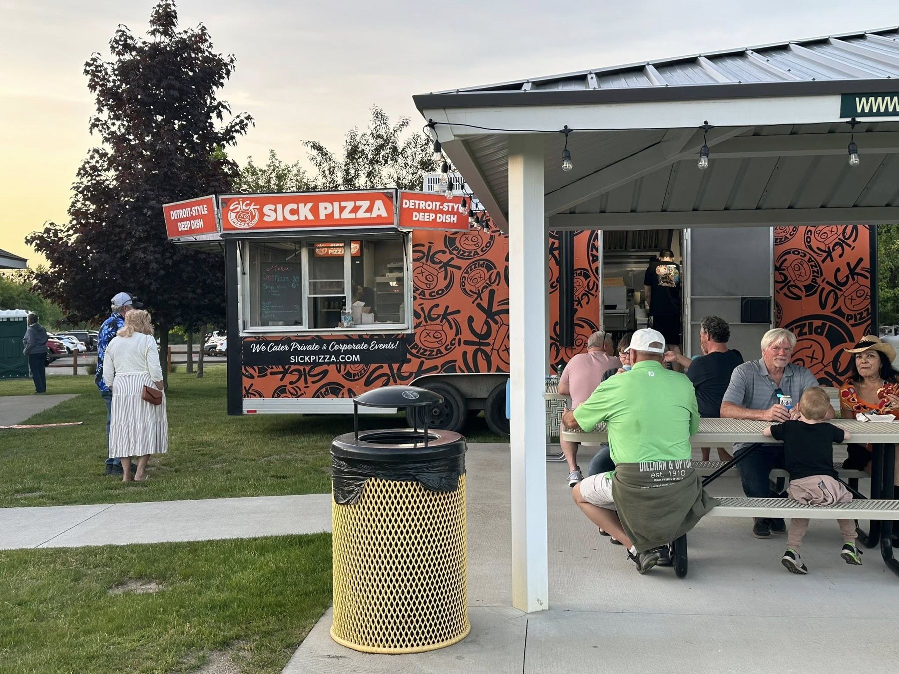 Guests eating pizza at picnic tables under the Wildwood pavilion, Sick Pizza trailer behind them